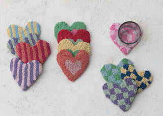 Colorful heart-shaped tufted coasters in stripe, check, and nested heart patterns displayed on white surface with glass.