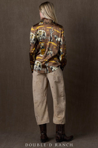 Woman wearing brown western-print satin blouse with cowboy scenes, paired with beige pants and dark cowboy boots.