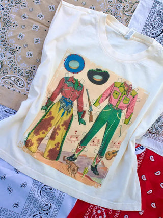 White graphic tee featuring vintage cowboy paper doll outfits in red, yellow, and green with Western accessories.