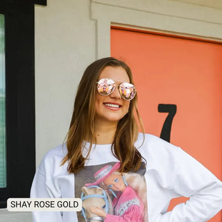 Rose gold aviator sunglasses with pink mirrored lenses worn by smiling woman in white sweatshirt.