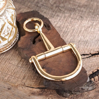 Shiny gold-tone horse bit keychain charm with D-ring and loop, displayed on rustic dark wood surface.
