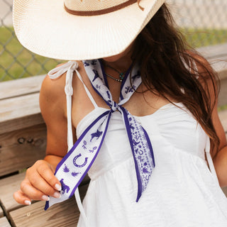 White and purple western-print twilly scarf tied around neck, featuring horseshoes, horses, and floral motifs.