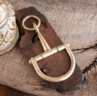 Worn gold-tone horse bit keychain charm with D-ring shackle and circular clasp on rustic wood surface.