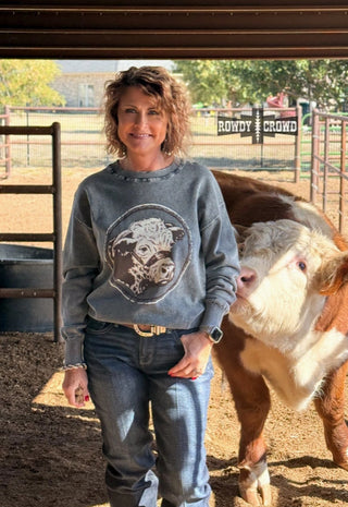 Woman wearing gray distressed sweatshirt featuring Hereford cow graphic, paired with jeans, standing beside real Hereford cal