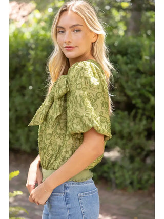 Olive green burnout floral top with large front bow detail and puff sleeves, paired with blue jeans.