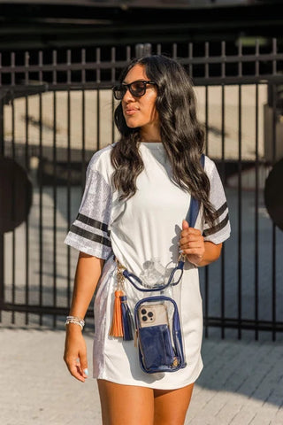 Woman wearing white dress carrying navy-trimmed clear stadium crossbody bag with phone pocket and tassel detail.