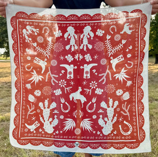 Red bandana with white Western-themed folk art print featuring cacti, horseshoes, longhorns, cowboys, and floral motifs.
