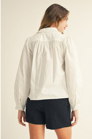 White long sleeve button-down shirt with ruffle collar and cuffs, gathered back detail, worn with navy shorts.