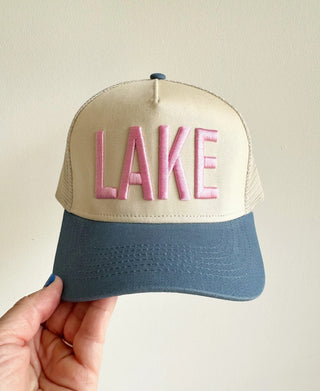 Beige and blue trucker hat with pink embroidered "LAKE" letters, held by hand against white background.