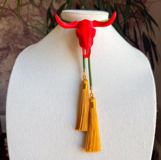Red longhorn skull bolo tie with olive green cord and dangling golden yellow tassel pendants.