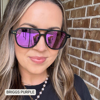 Woman wearing Briggs black aviator sunglasses with purple mirrored polarized lenses against brick wall.