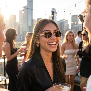 Woman wearing black oval-frame sunglasses with gold accents at a rooftop party, smiling in golden hour light.