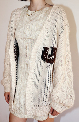 Cream open-knit oversized cardigan with brown horse print detail, layered over cable-knit sweater dress.