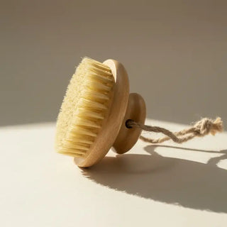 Round wooden dry body brush with natural cream-colored sisal bristles and jute hanging loop, on neutral background.