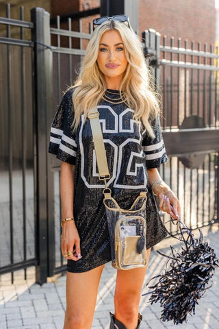 Clear stadium bag with gold trim crossbody strap, worn by blonde woman in sequin jersey dress holding black pom-poms.