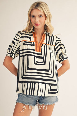 Woman wearing cream and black geometric print short sleeve top with orange V-neck contrast trim accent.