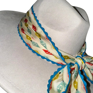 Cream twilly scarf with colorful diamond pattern and blue rickrack trim, tied around white hat display.