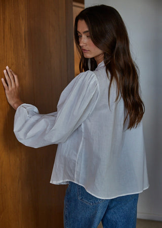 White woven cotton button-down blouse with voluminous balloon sleeves and ruffled collar, paired with blue jeans.