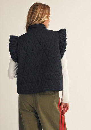 Black quilted vest with ruffled cap sleeves, worn over white long-sleeve top with olive pants, back view.