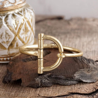 Gold equestrian horsebit hook bracelet displayed on rustic driftwood with decorative glass jar background.