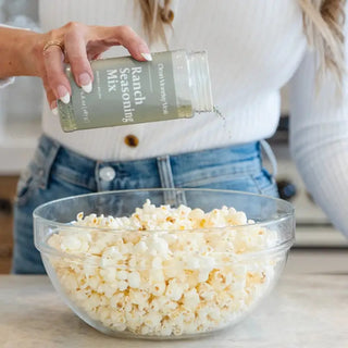 Woman in white top sprinkling Ranch Seasoning Mix from sage green bottle onto bowl of popcorn.