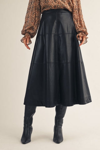 Black vegan leather tiered A-line midi skirt with paneled seam details, styled with knee-high boots.