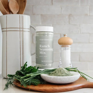 Clean Monday Meals Ranch Seasoning Mix jar on wooden board with fresh herbs, salt grinder, and green seasoning powder.
