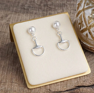 Silver drop earrings featuring round white pearl studs and horse bit pendants on cream jewelry display.