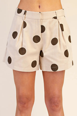 Cream high-waist pleated shorts with large dark brown polka dots, featuring belt loops and a tailored fit.