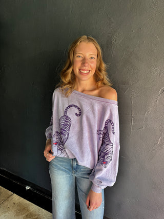 Lavender off-shoulder sweatshirt featuring purple tiger graphics, paired with light wash jeans.