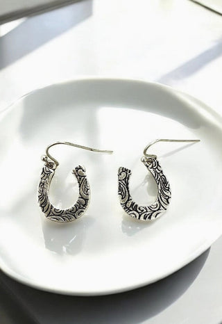 Antique silver horseshoe dangle earrings with intricate floral scroll engraving, displayed on white ceramic dish.