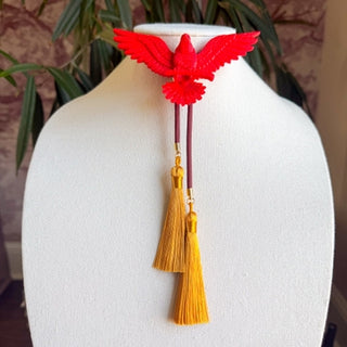 Red eagle-shaped bolo tie with maroon cord and two golden yellow tassel pendants displayed on white jewelry bust.
