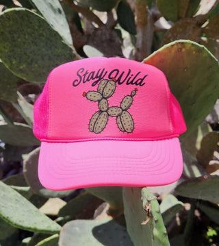 Neon pink trucker hat with "Stay Wild" text and cactus balloon dog graphic, displayed outdoors near cacti.