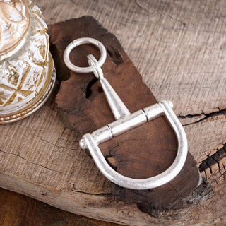 Silver-tone horse bit keychain charm with D-ring design displayed on rustic dark wood surface.