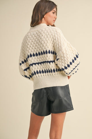 Cream chunky knit sweater with navy blue stripe and diamond pattern, paired with black leather shorts.