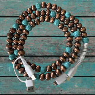 Copper and turquoise beaded charging cable coiled on rustic teal wood surface with USB and Lightning connectors visible.