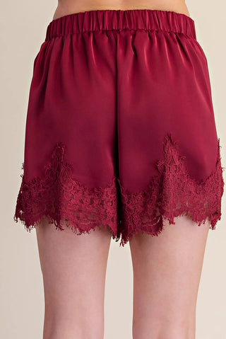 Burgundy satin shorts with elegant floral lace trim detail along scalloped hemline and elastic waistband.