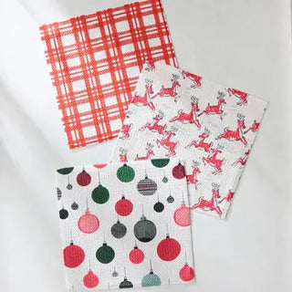 Three festive Christmas dishcloths featuring red plaid, prancing reindeer, and colorful holiday ornament patterns.