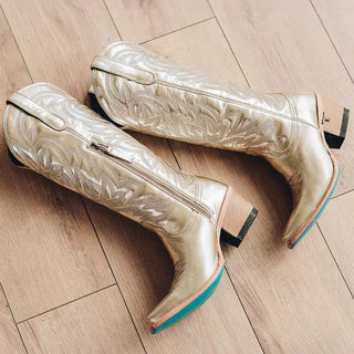 Champagne metallic leather cowboy boots with embroidered detailing, block heel, and turquoise sole on wood floor.