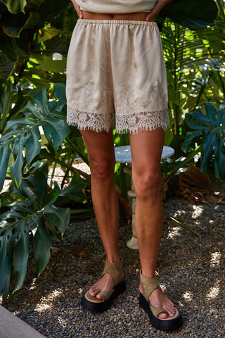 Beige satin shorts with floral lace trim and fringe hem, paired with olive platform sandals.