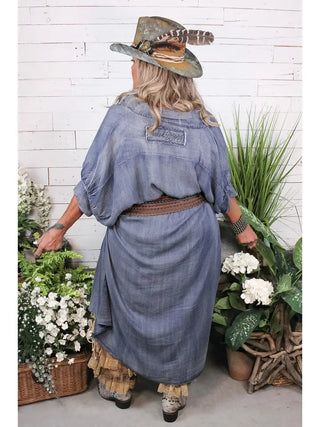 Woman wearing a long chambray blue denim Morning Meadow Dress with ruffled hem, brown studded belt, and cowboy hat.