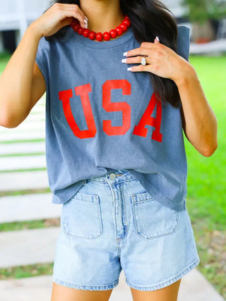 Navy blue sleeveless USA muscle tank with bold red lettering, styled with denim shorts and red beaded necklace.