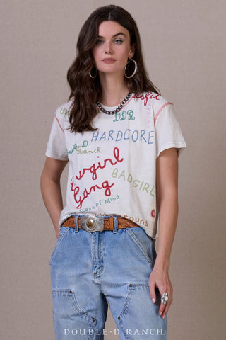 White graphic tee with colorful embroidered text including "Hardcore" and "Bad Girl," paired with blue jeans and western belt