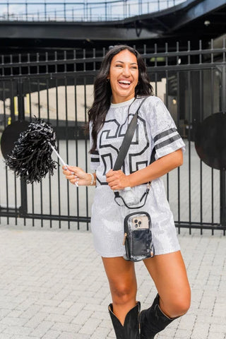 Clear stadium crossbody bag with black trim, worn by smiling woman in silver sequin outfit with black pom-pom.