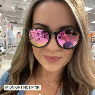 Woman wearing black horn-rimmed polarized sunglasses with hot pink mirrored lenses in a retail store.