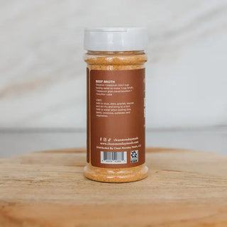 Brown glass shaker jar of Clean Monday Meals Beef Bouillon seasoning with white lid on wooden surface.