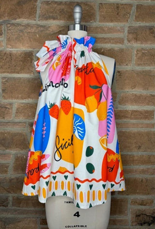 Colorful sleeveless ruffle tie-neck top featuring fruit print and Italian text on white fabric.
