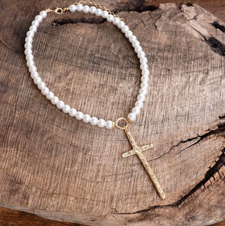 Gold primitive cross pendant on white pearl beaded necklace displayed on rustic wood surface.