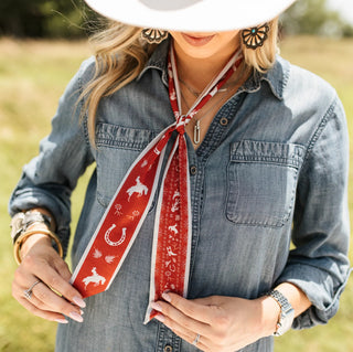 Red western-print twilly scarf tied at neckline, featuring cowboys, horseshoes, and bandana motifs with cream border.