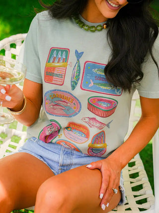 Sage green graphic tee featuring colorful vintage sardine can illustrations in pink, teal, and gold tones.